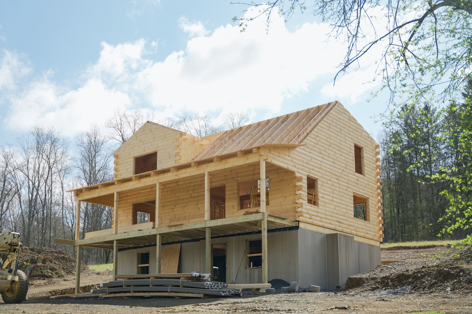 A partially completed white pine log home under construction, with log walls raised and roof rafters in place over a poured foundation