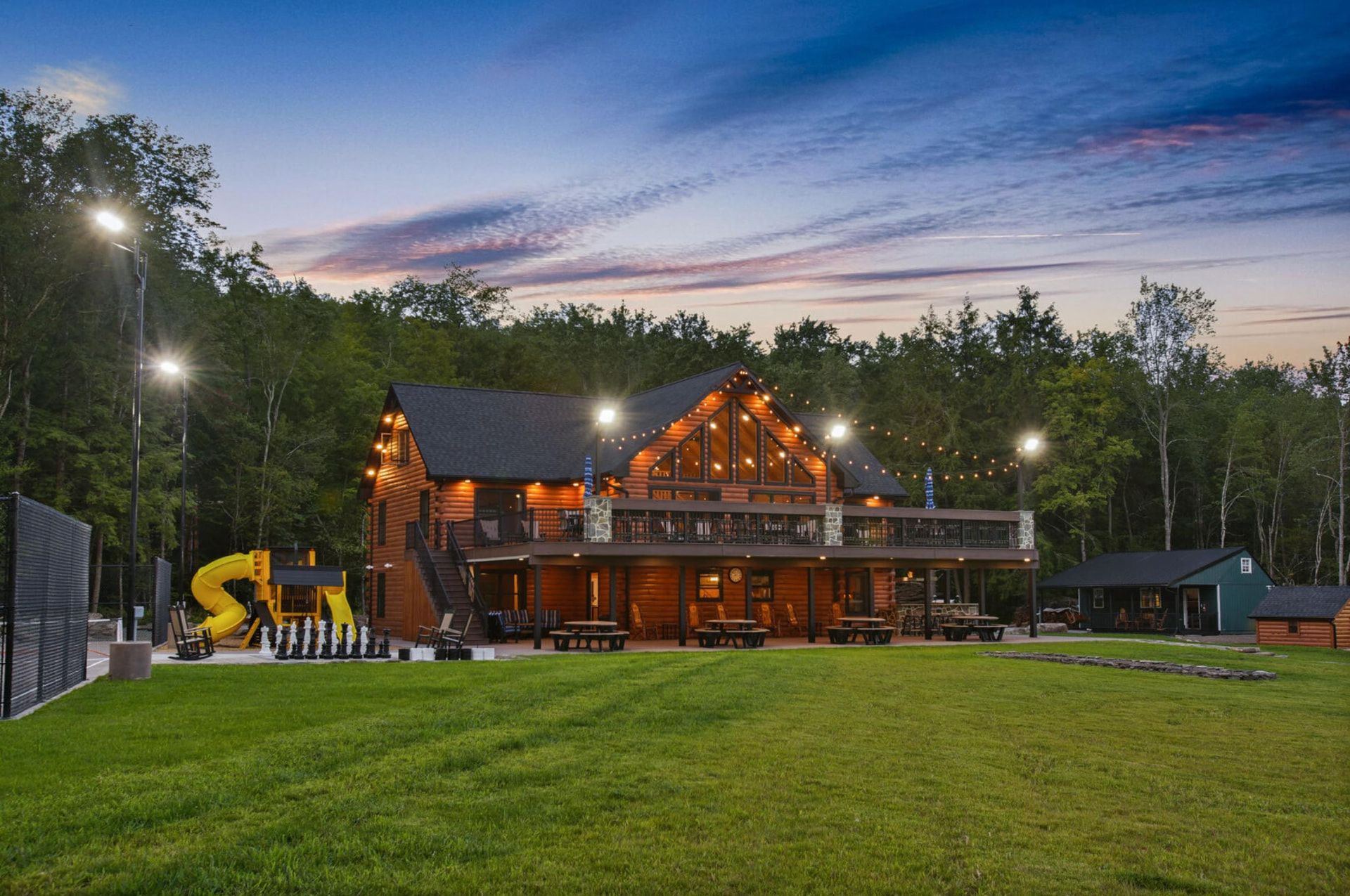 A&A Cabin log lodge at dusk with string lights, wraparound deck and playground